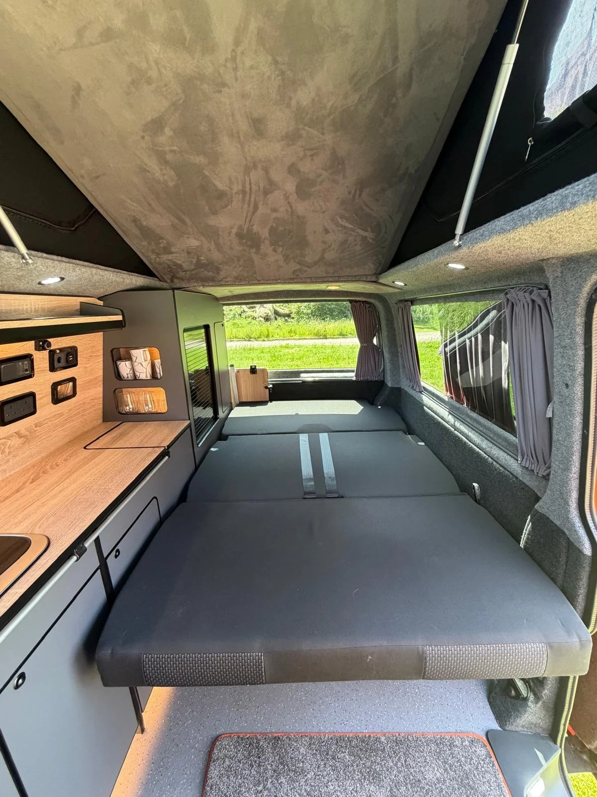 The interior of a camper van with a fold-out bed, curtains on the windows, and a wooden countertop with electrical outlets and switches.