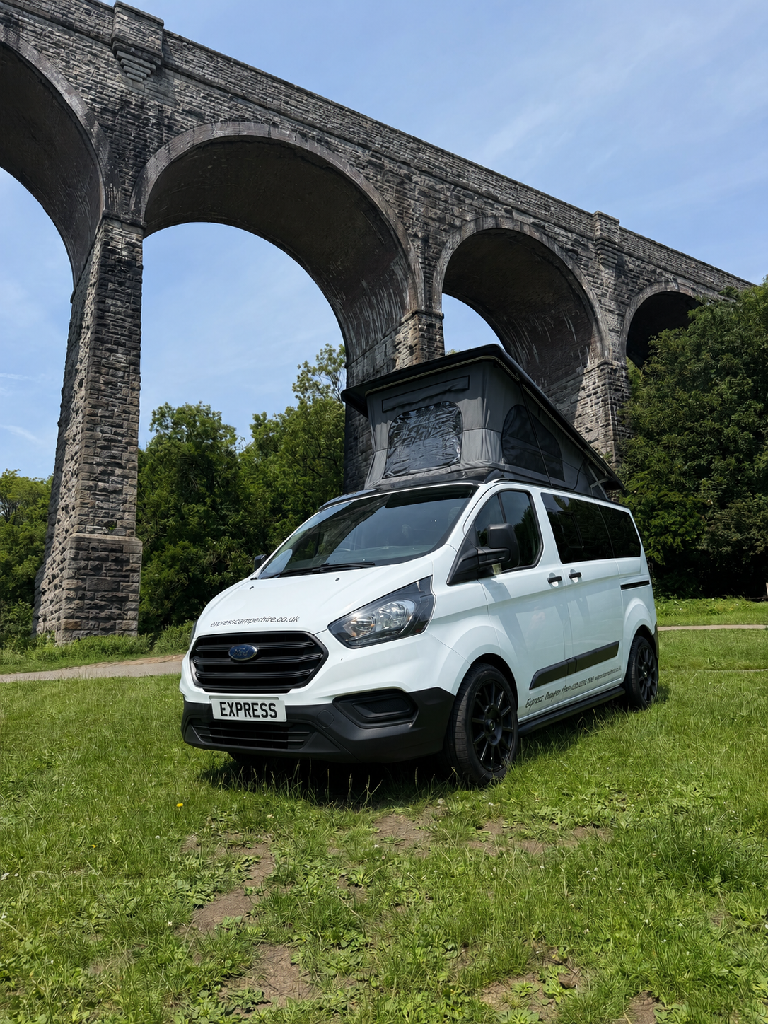 White camper van with a pop-up roof parked on grass near a stone arch bridge.