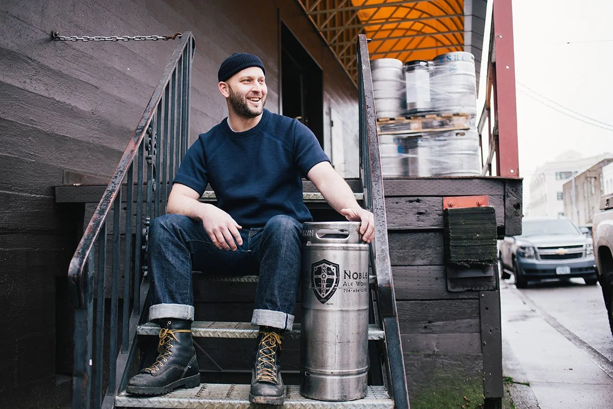 Robby Roda sitting on outdoor stairs next to a beer keg, smiling, wearing a navy blue T-shirt, jeans, black boots, and a black beanie, with a stack of beer kegs in the background.