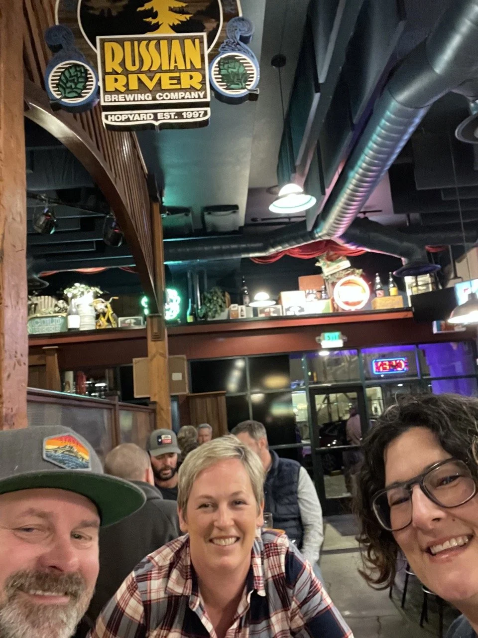 A group of three smiling people taking a selfie inside a lively bar or restaurant, with a Russian River Brewing Company sign hanging from the ceiling.