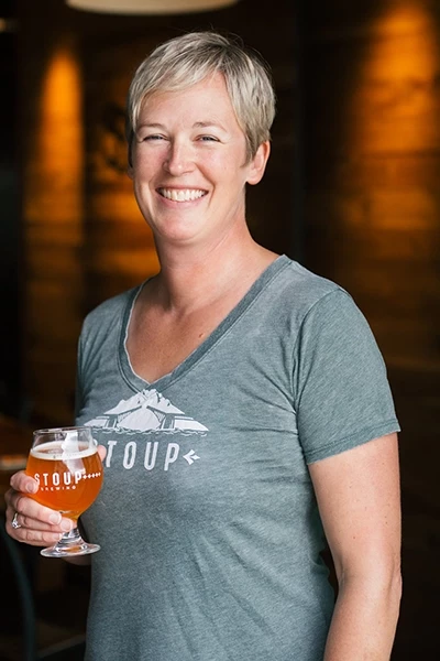 Robyn Schumacher, with short blonde hair holding a glass of beer, standing in a dimly lit bar or brewery.