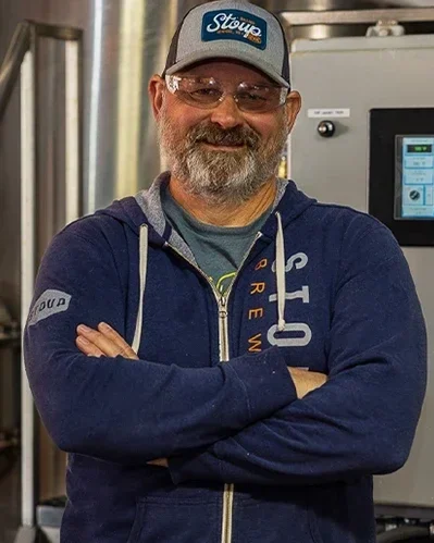 Brad Benson, wearing a blue hoodie and a trucker hat, standing with arms crossed in front of industrial equipment.