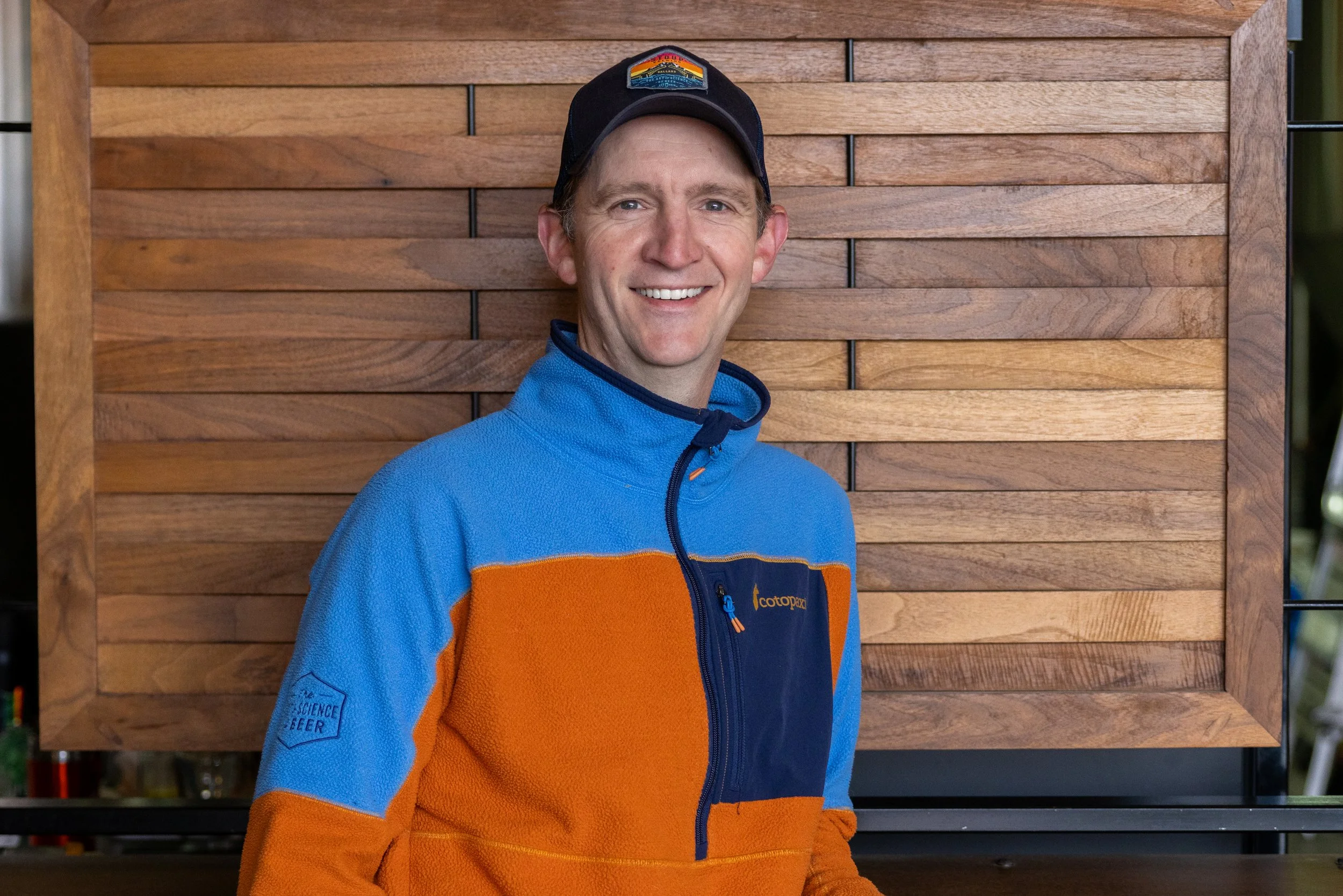 Adam Lasowska smiling and wearing a black cap and a blue and orange jacket standing in front of a wooden wall.