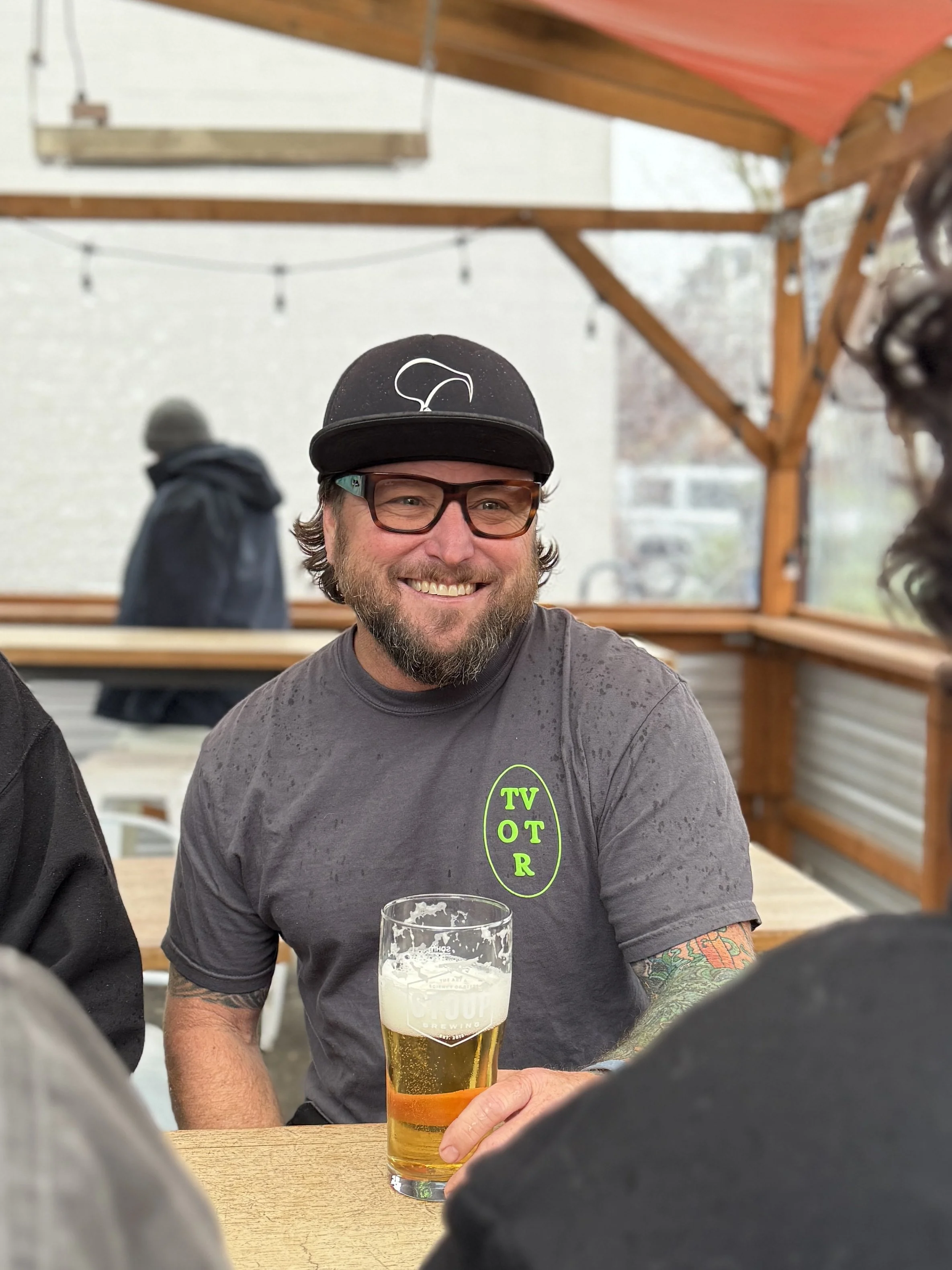Jason Bass, with glasses and a black baseball cap is sitting at a wooden table with a glass of beer in front of him. The background shows a covered outdoor area with string lights and a person in a hoodie walking by.