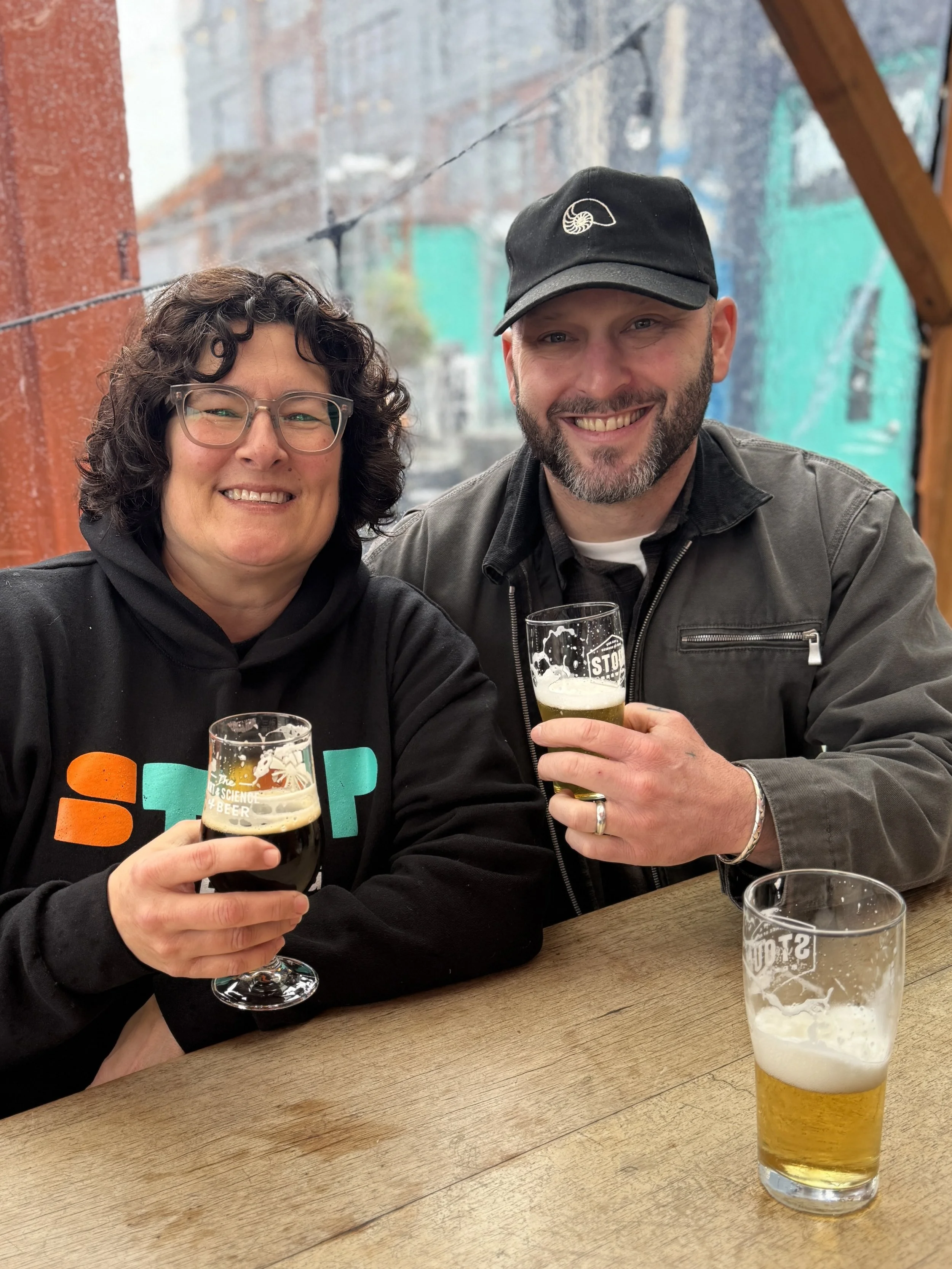 Lara Zahaba and Robby Roda are holding glasses of beer. They are sitting at a wooden table in a restaurant or bar, with a blurred background of window and brick wall.