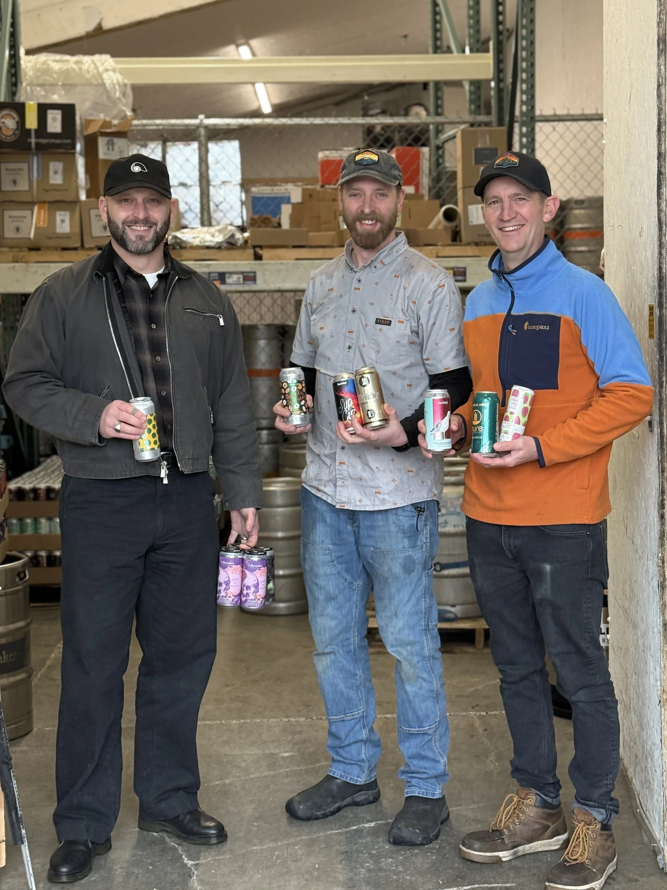Three men in hats stand in front of an open warehouse door, holding cans of beer.