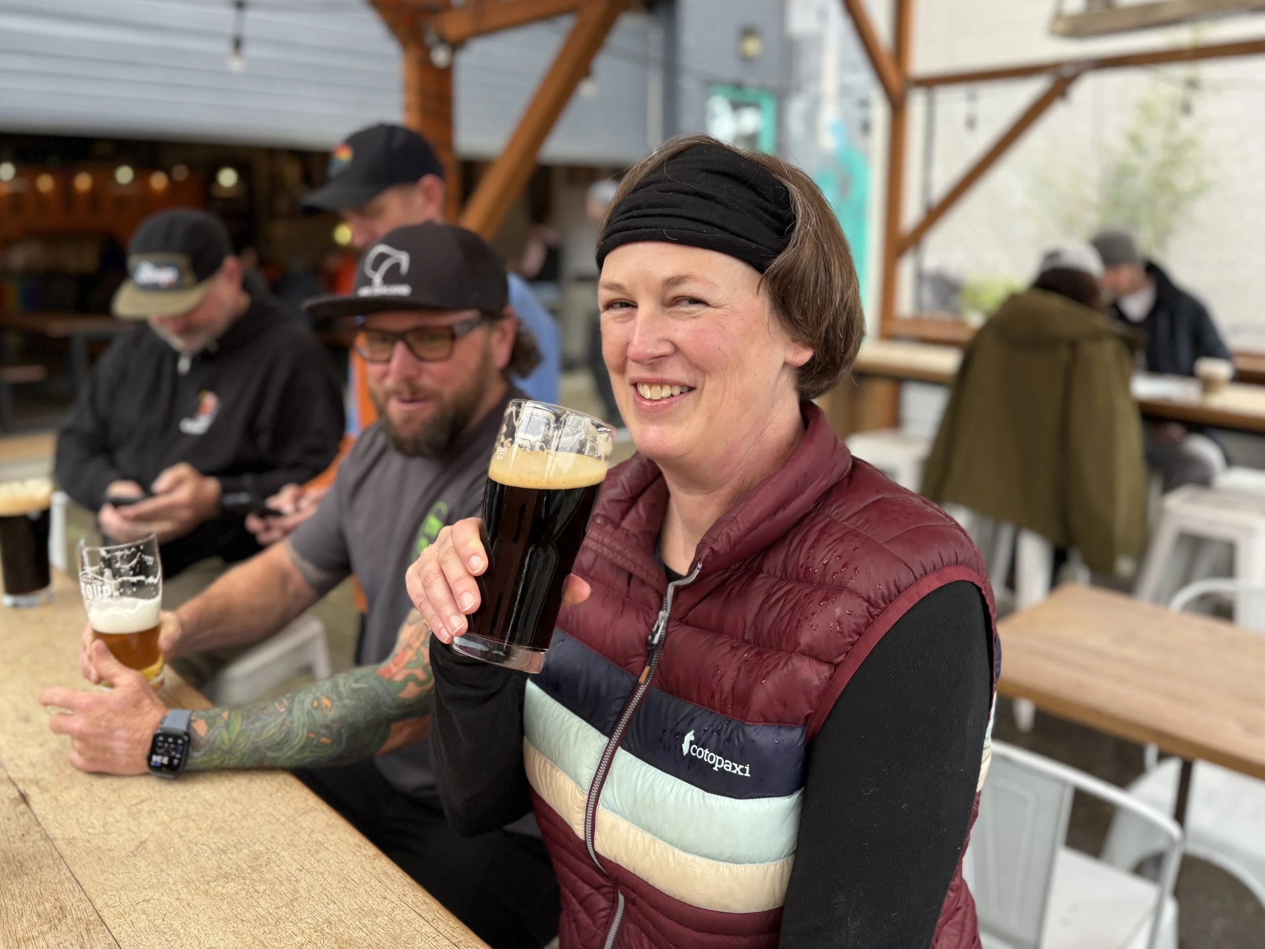 A woman smiling and holding a glass of dark beer at a bar or brewery, with other people sitting at the same table.