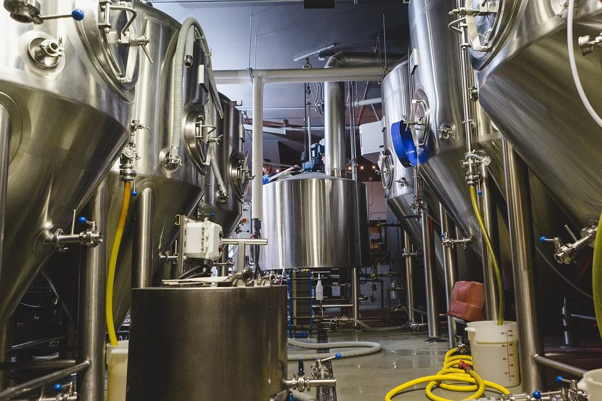 Industrial brewery equipment made of stainless steel, including large fermentation tanks, pipes, and hoses.