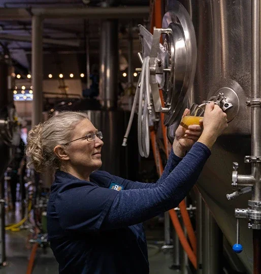 A woman with glasses, blonde hair in a bun, wearing a navy blue jacket working in the brewhouse.