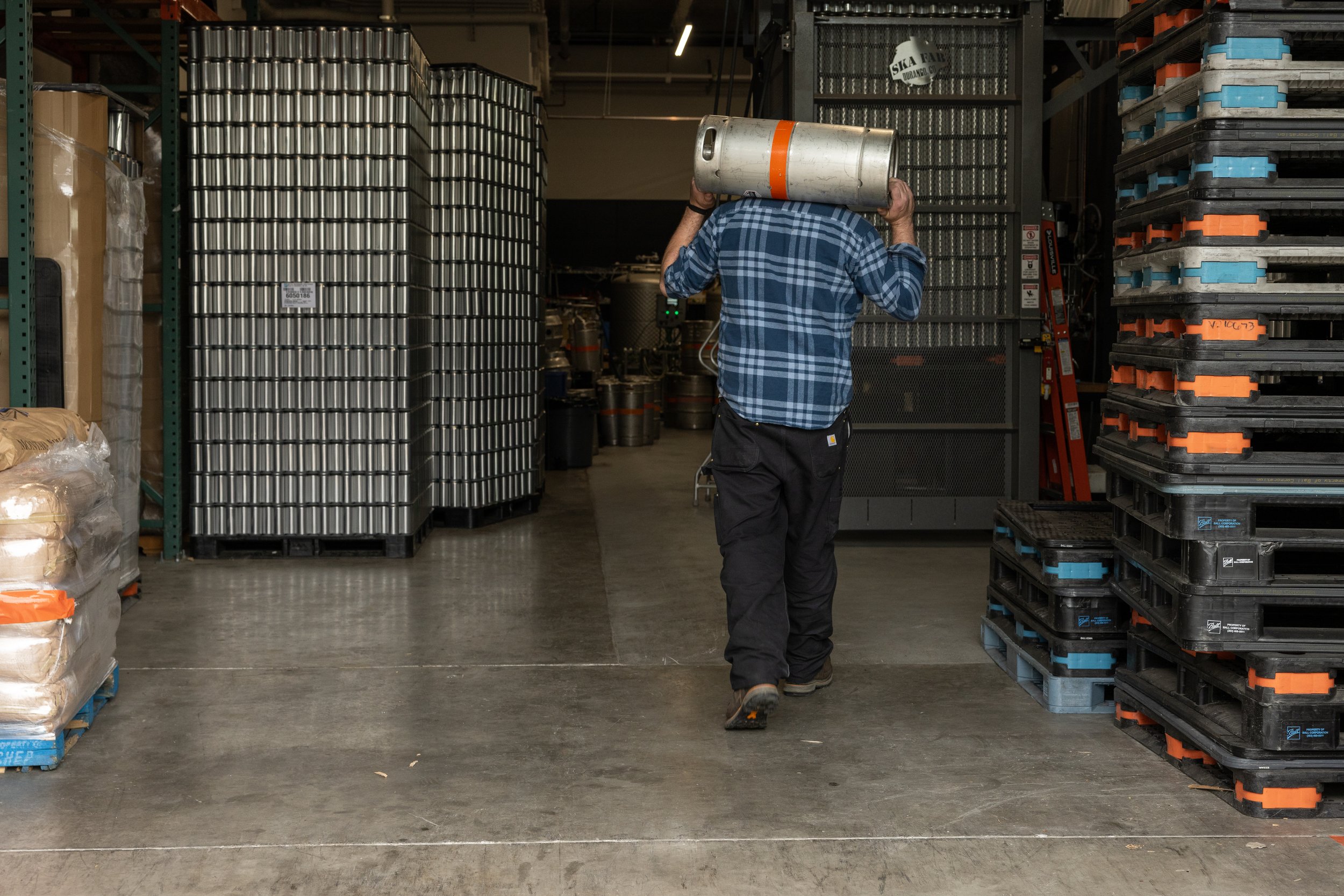 A person carrying a large metal keg in a warehouse, surrounded by stacked pallets.