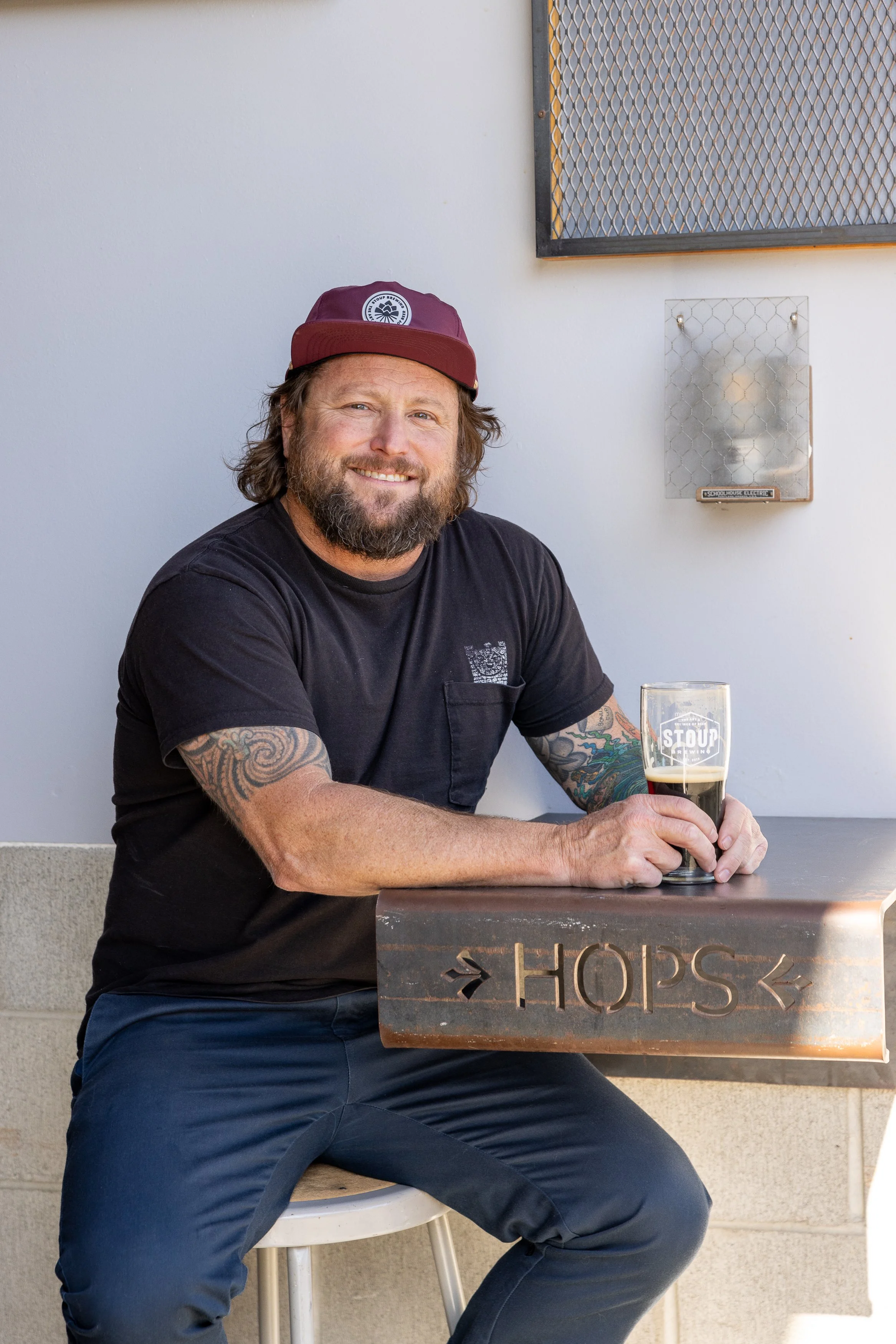 Jason Bass, with a beard and tattoos, wearing a maroon cap and black t-shirt, smiling and sitting at a Stoup Brewing with glass of dark beer.