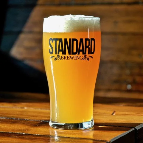 A glass of beer with foam on top, branded with 'Standard Brewing' on the glass, placed on a wooden table with a blurred wooden background.