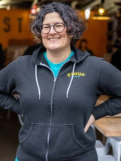 Lara Zahaba, with curly hair, glasses, smiling, wearing a black hoodie with a yellow logo, standing indoors in a restaurant or a cafe.