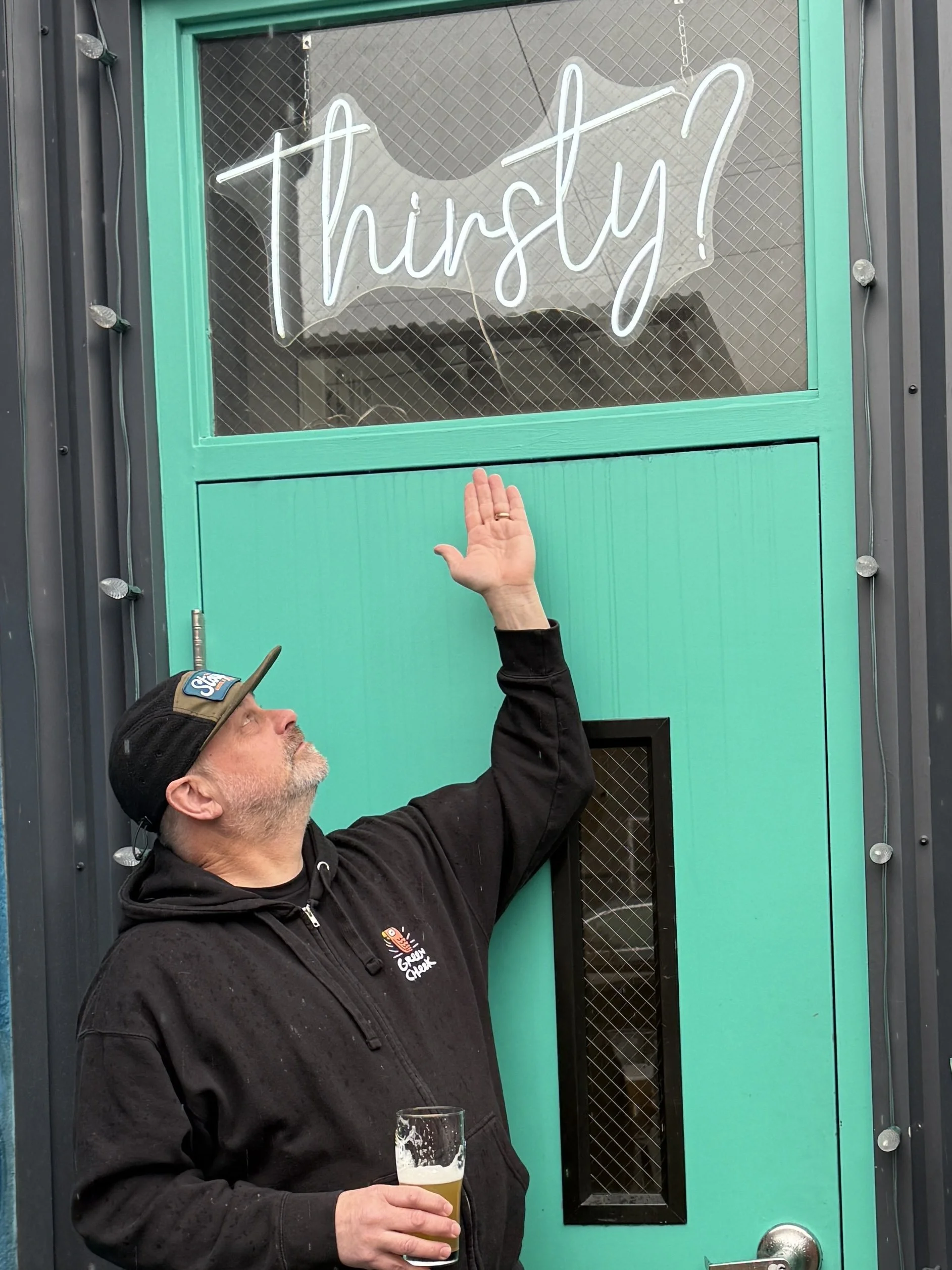 Brad Benson wearing a black hoodie and a black cap with a beer in his hand is reaching out towards a glowing white script sign that reads 'thirsty!' on a teal door.