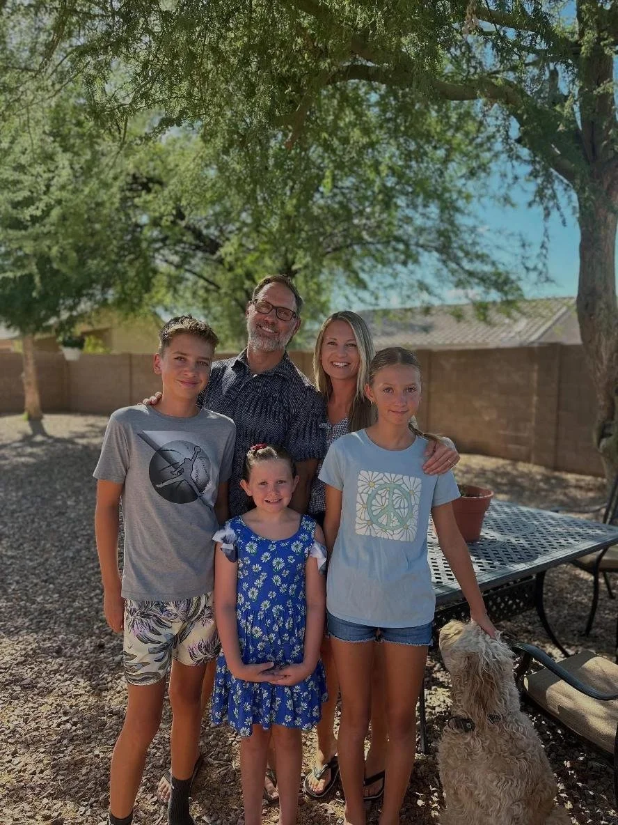 A family of five standing outdoors under a large tree, smiling, with a brick wall in the background. Two children are girls in blue dresses, and a boy in a gray t-shirt and shorts. Two adults, a man and woman, are in the center. A dog is sitting next to one of the girls, and there is a metal outdoor table with a potted plant on it.