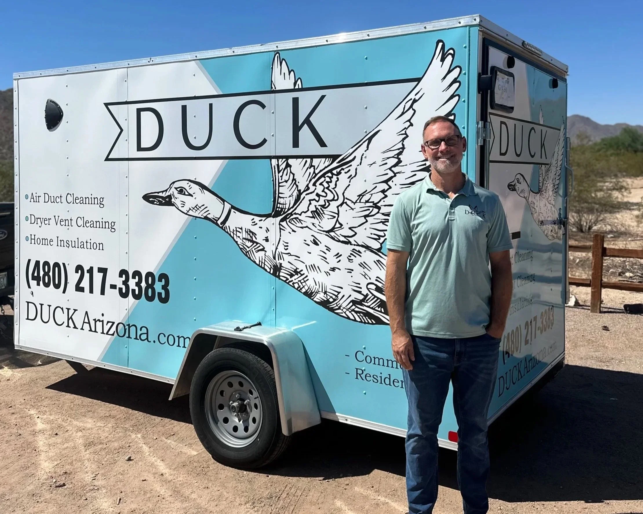A man wearing glasses and a light green polo shirt with a logo, standing beside a blue mobile service trailer with a duck logo and the word 'DUCK' on it. The trailer offers services like air duct cleaning, dryer vent cleaning, and home insulation, and is set against a desert landscape with hills and a clear blue sky.