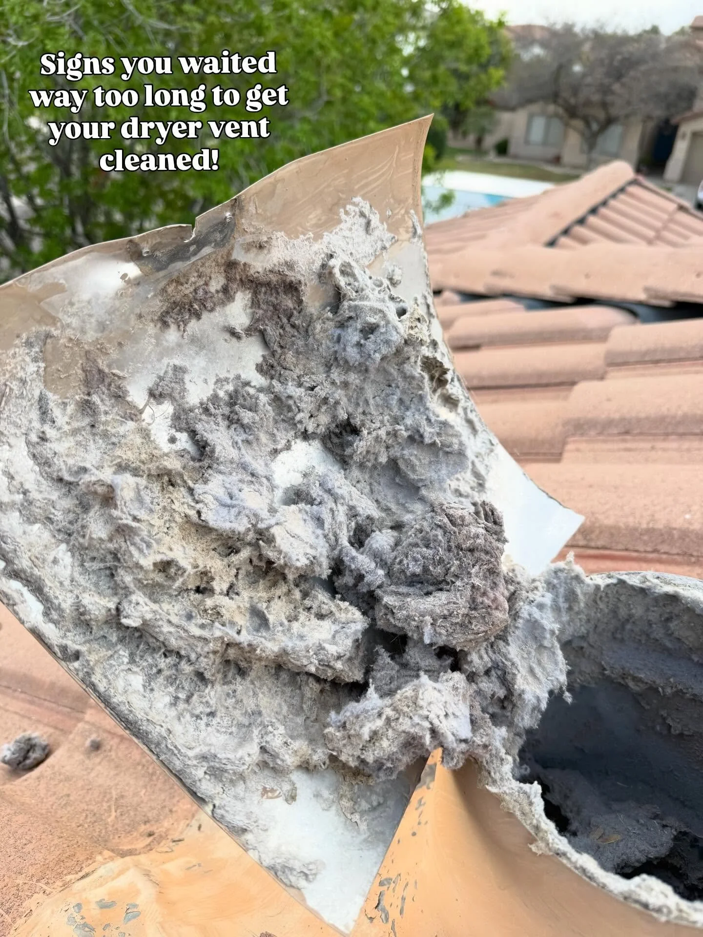 Close-up of a damaged dryer vent covered in lint and debris with a roof and trees in the background.
