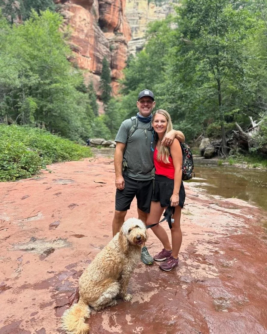 A smiling couple with a dog on a leash standing on a wet reddish rock trail beside a river, surrounded by lush green trees and red canyon cliffs.