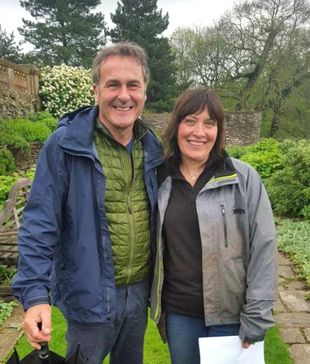 A man and woman smiling outdoors in a garden with trees and shrubs, both wearing jackets.