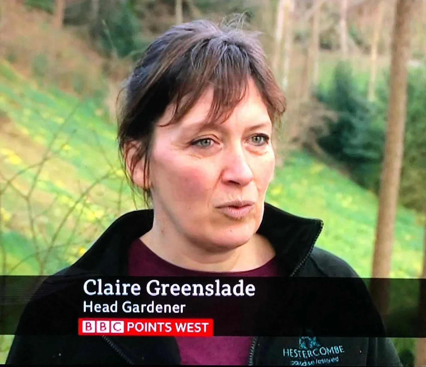 A woman with short brown hair and blue eyes speaking outdoors with trees and green foliage in the background. She is wearing a black jacket with a logo on it. The text on screen identifies her as Claire Greenslade, Head Gardener, on BBC Points West.