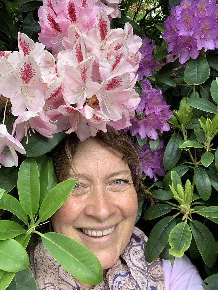 Claire Greenslade smiling among pink and purple rhododendron flowers and green leaves.