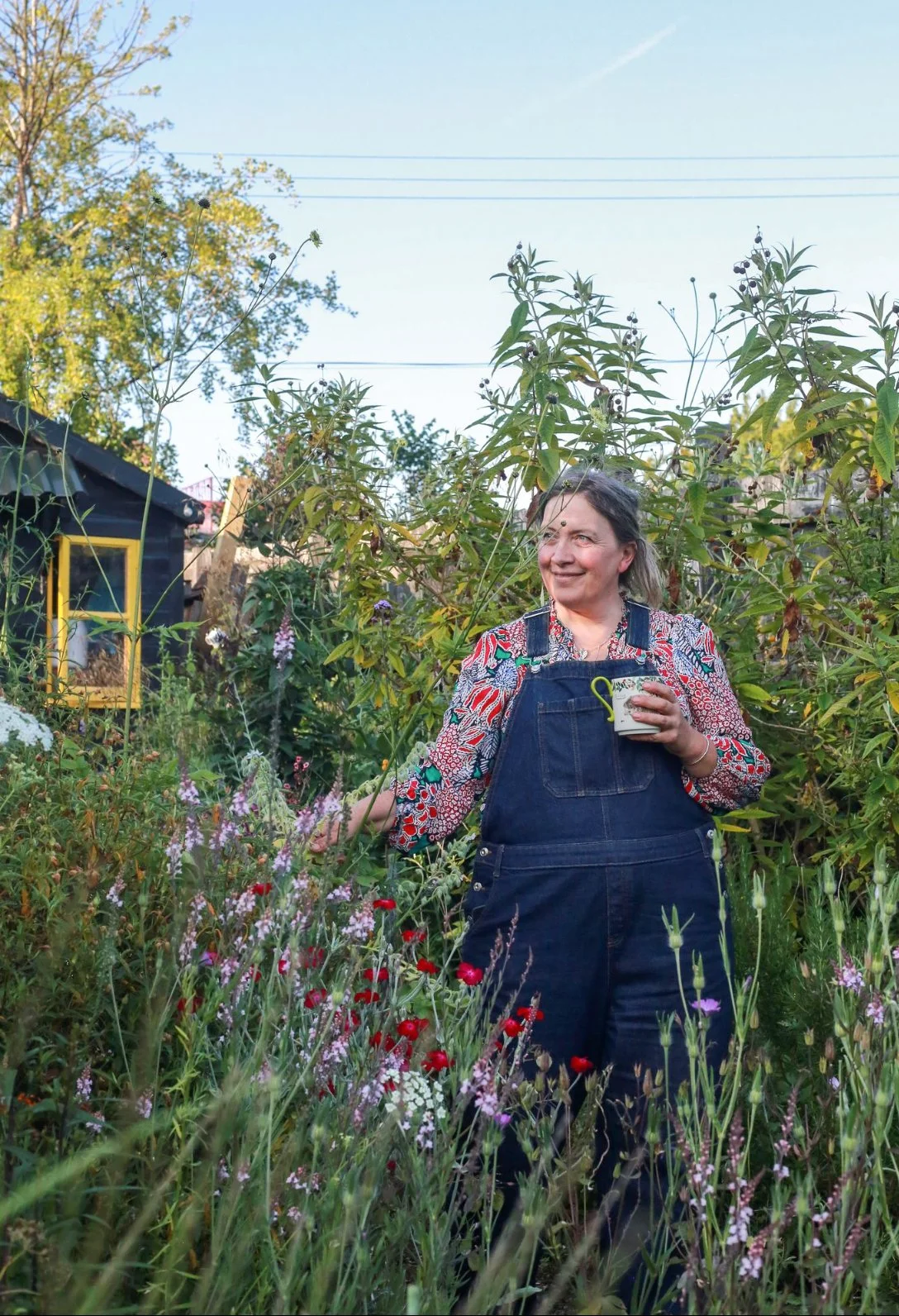Claire Greenslade holding a cup of tea, standing in a lush garden with various flowering plants and shrubs, smiling and enjoying the outdoor setting during daylight.