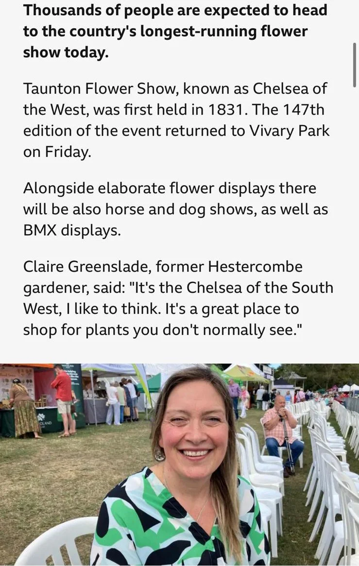 A smiling woman sitting outdoors at a festival with white chairs and a crowd in the background, and a large sign mentioning the Taunton Flower Show.
