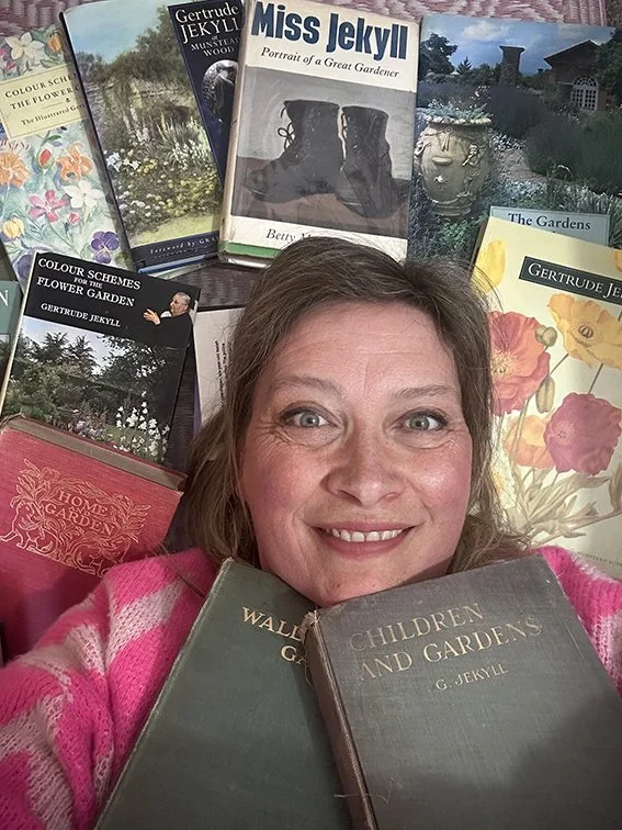 Claire Greenslade laying on the floor surrounded by gardening books that show she is an expert in the work of Gertrude Jekyll She is smiling and wearing a pink and gray striped sweater.