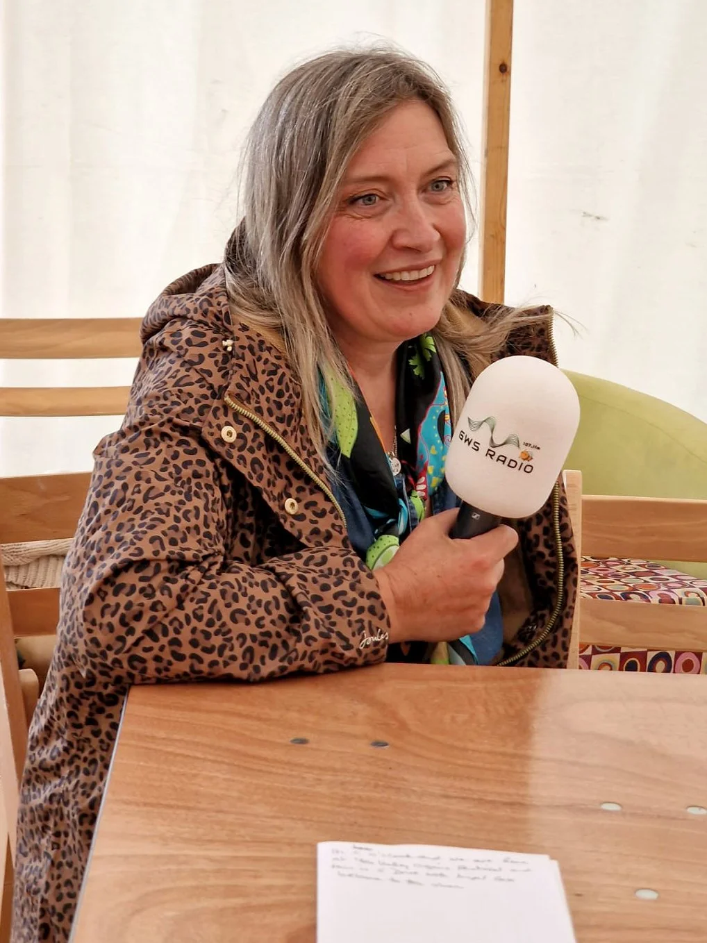 Woman with blonde hair smiling and holding a microphone labeled "GWS Radio" in a casual indoor setting.