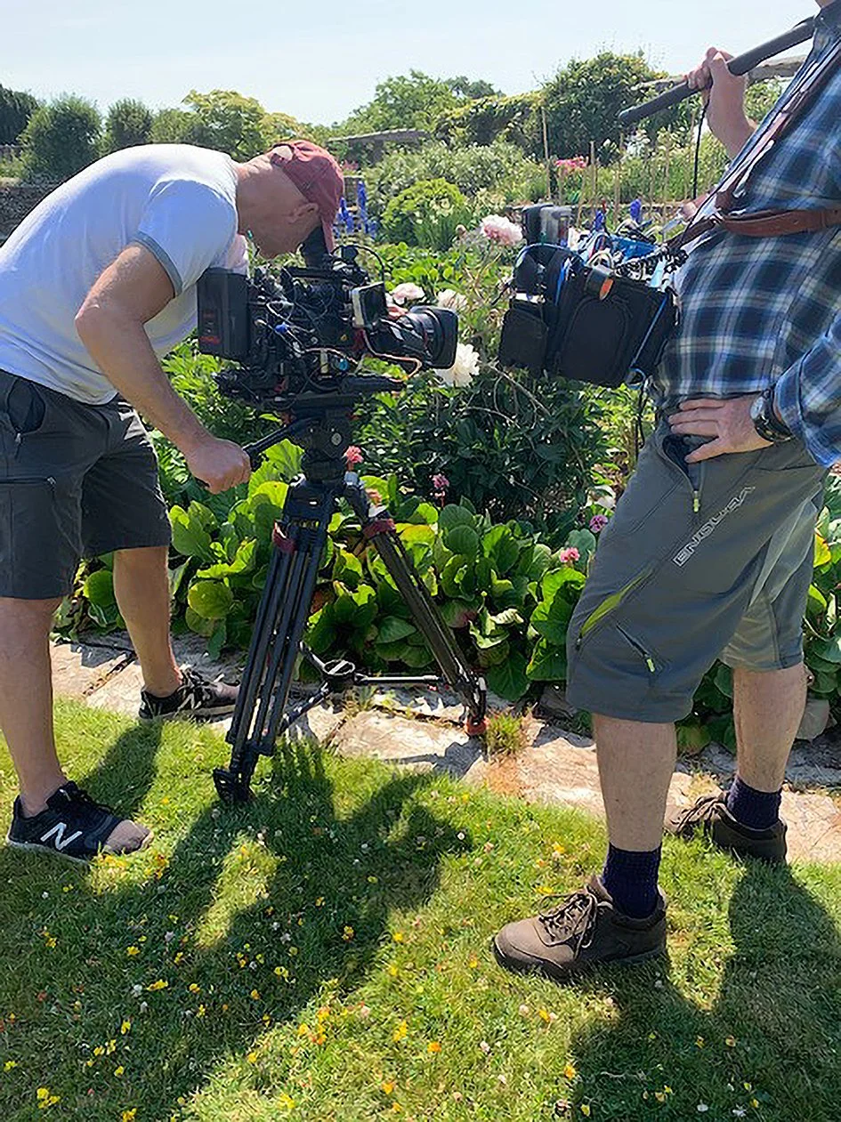Two videographers filming in a garden with green plants and flowers under a bright sunny sky.