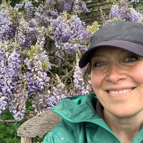 Claire Greenslade wearing a gray cap and green jacket smiling outdoors near purple wisteria flowers.