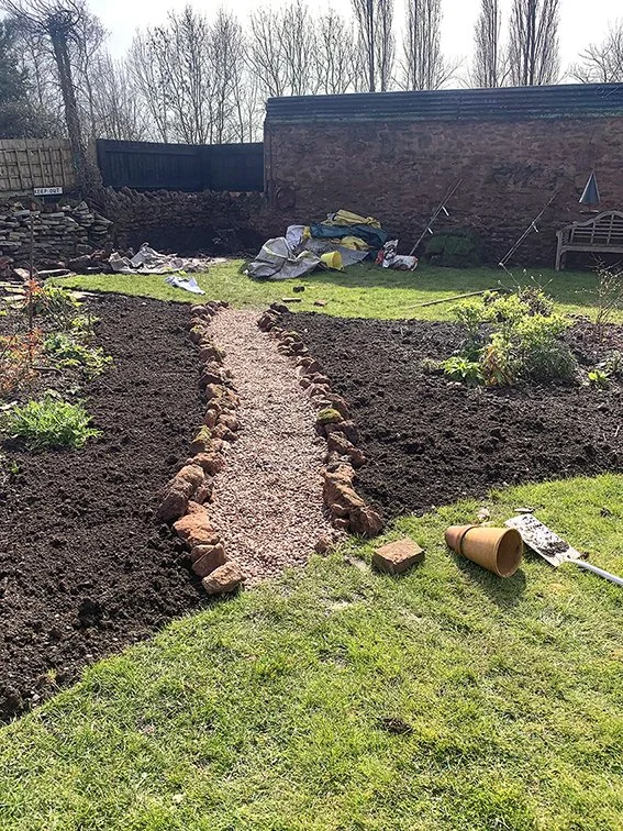 A garden with freshly tilled soil on both sides of a gravel path lined with small rocks, with gardening tools, a flower pot, and supplies in the background.