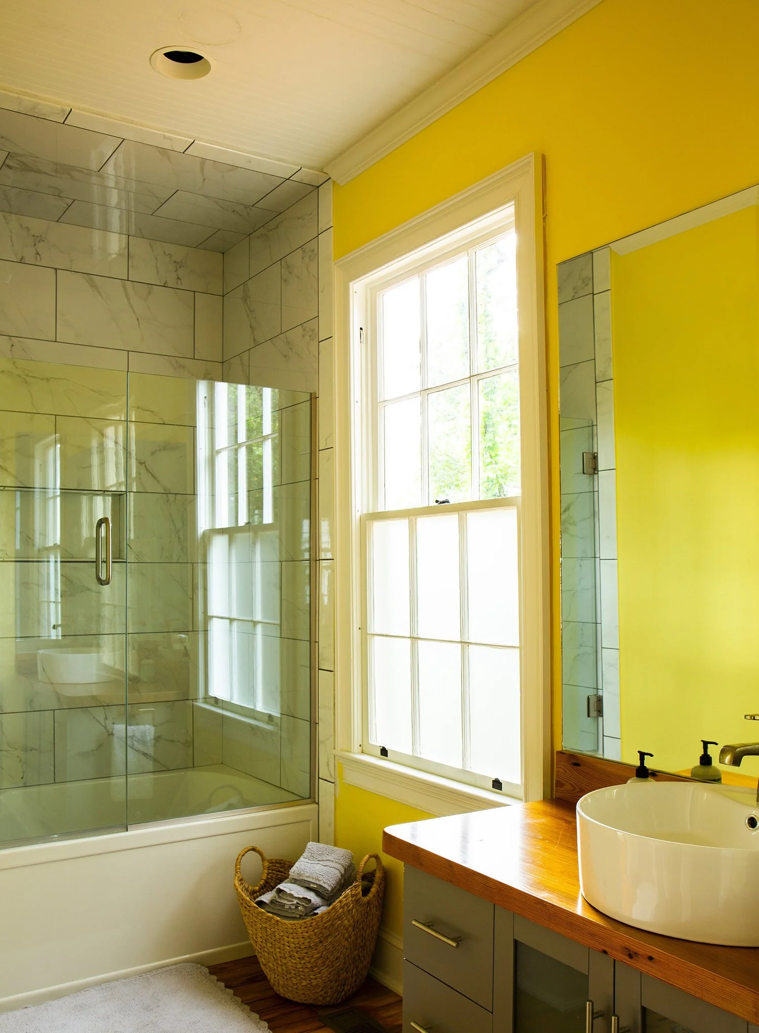 Bright bathroom with a shower stall enclosed by glass, a large window with lots of natural light, yellow walls, a wooden countertop with a white vessel sink, and a basket with rolled-up towels.