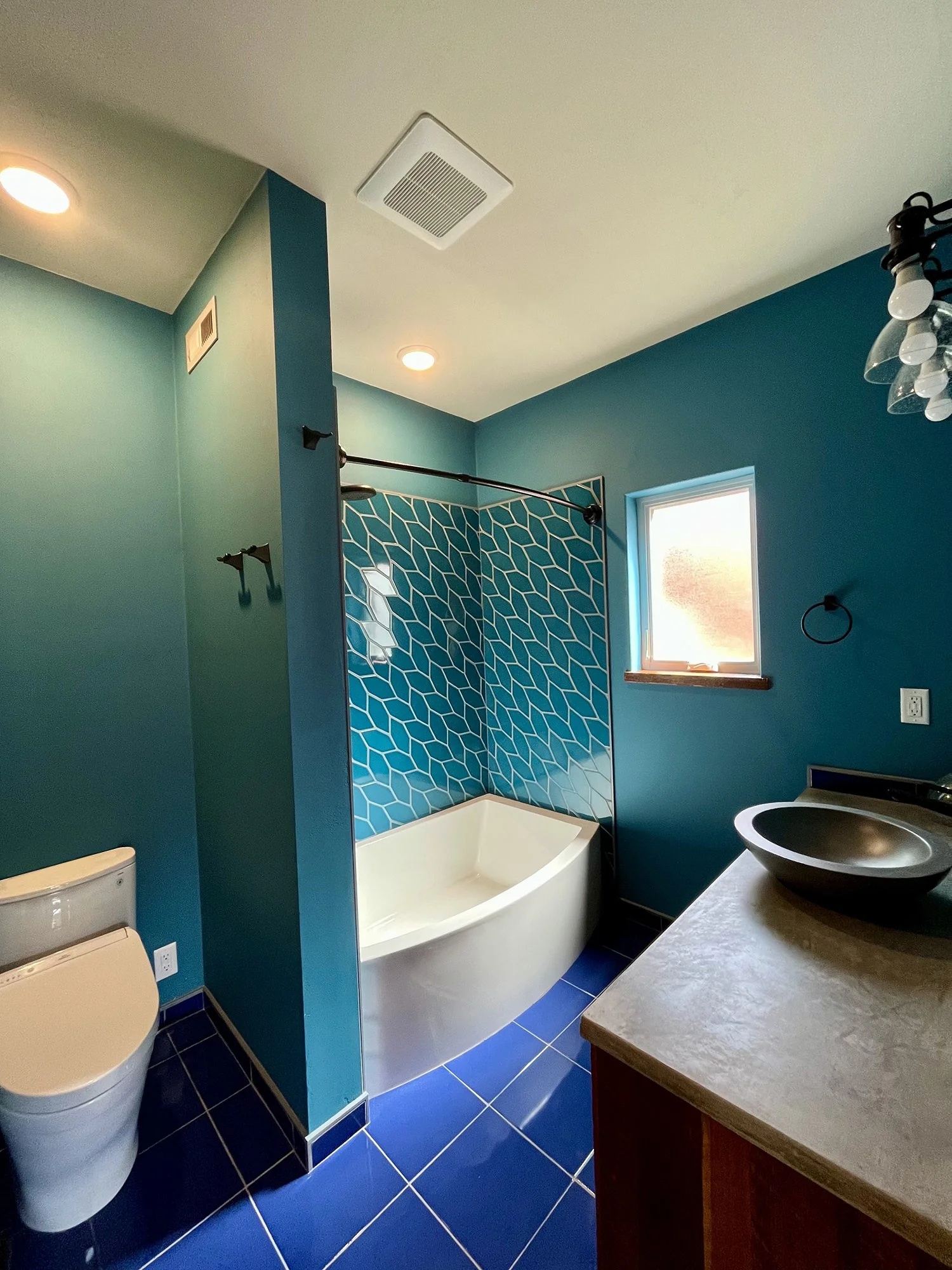 Modern bathroom with blue tiled floor, teal walls, a corner bathtub with a blue geometric tile accent wall, a small window, a floating sink with a round black basin, and a toilet.