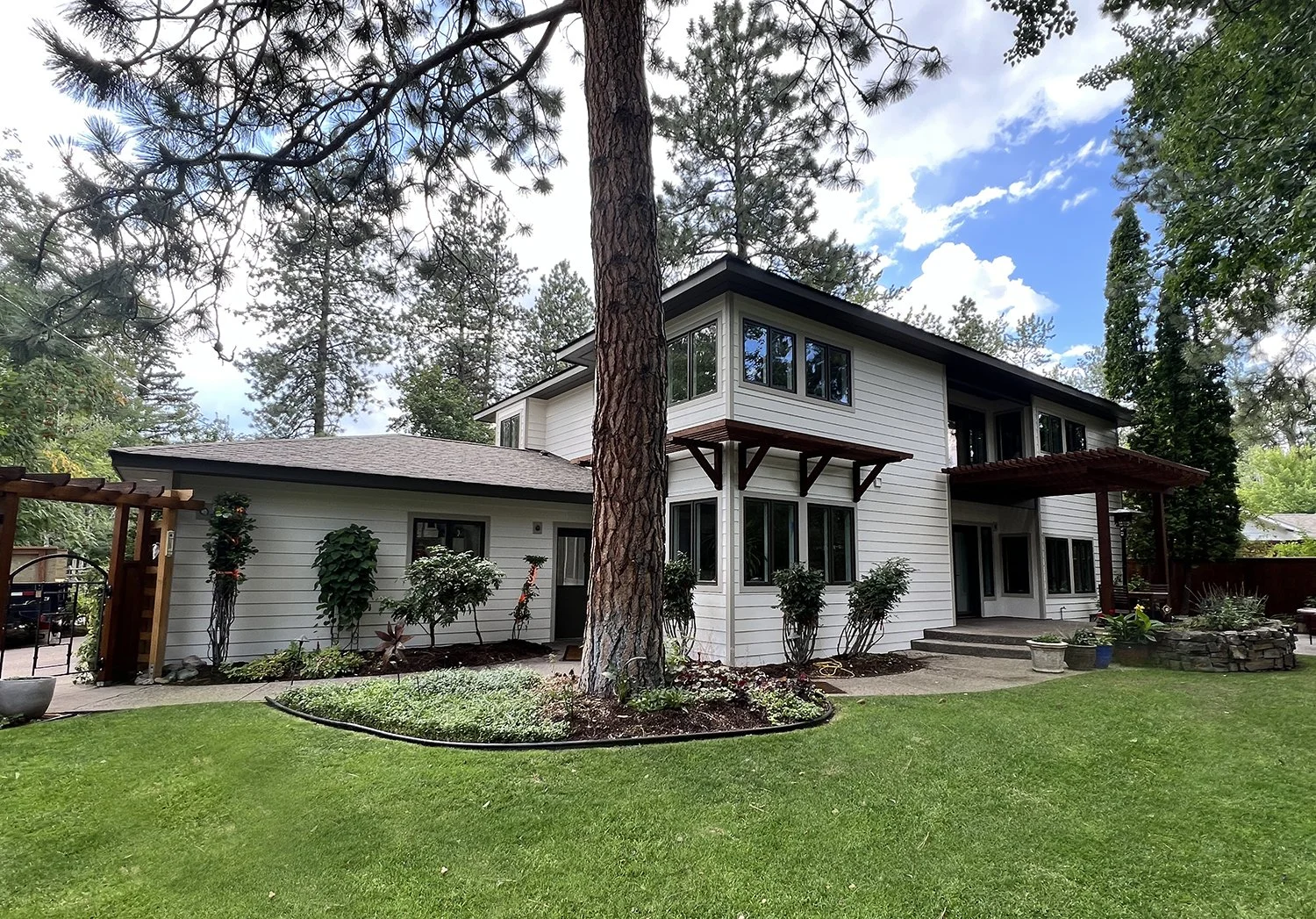 A two-story white house with black windows, surrounded by green trees and a well-maintained lawn, with a large pine tree in the front yard and steps leading to the entrance.