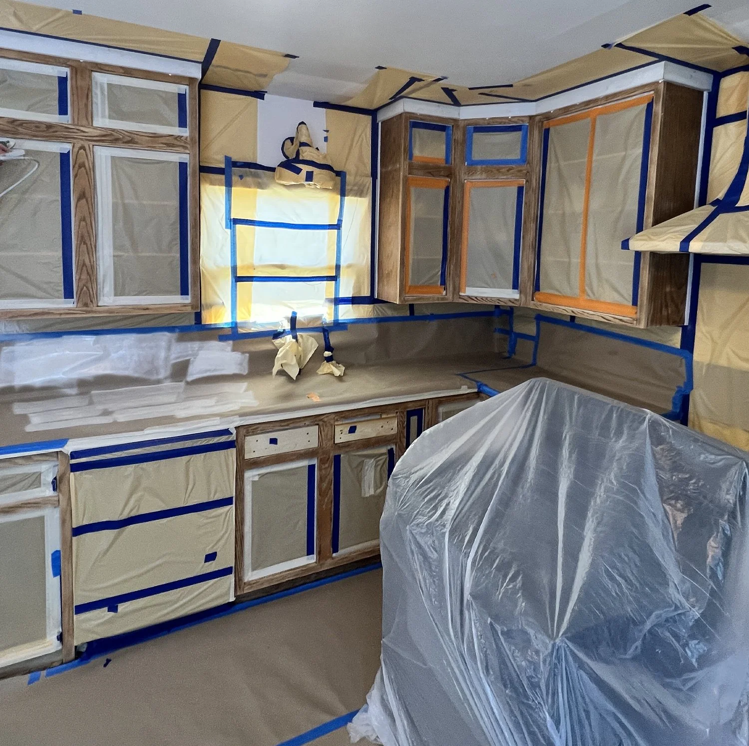 Kitchen cabinets under construction with protective plastic and painter's tape in a room, with a covered object in the foreground.