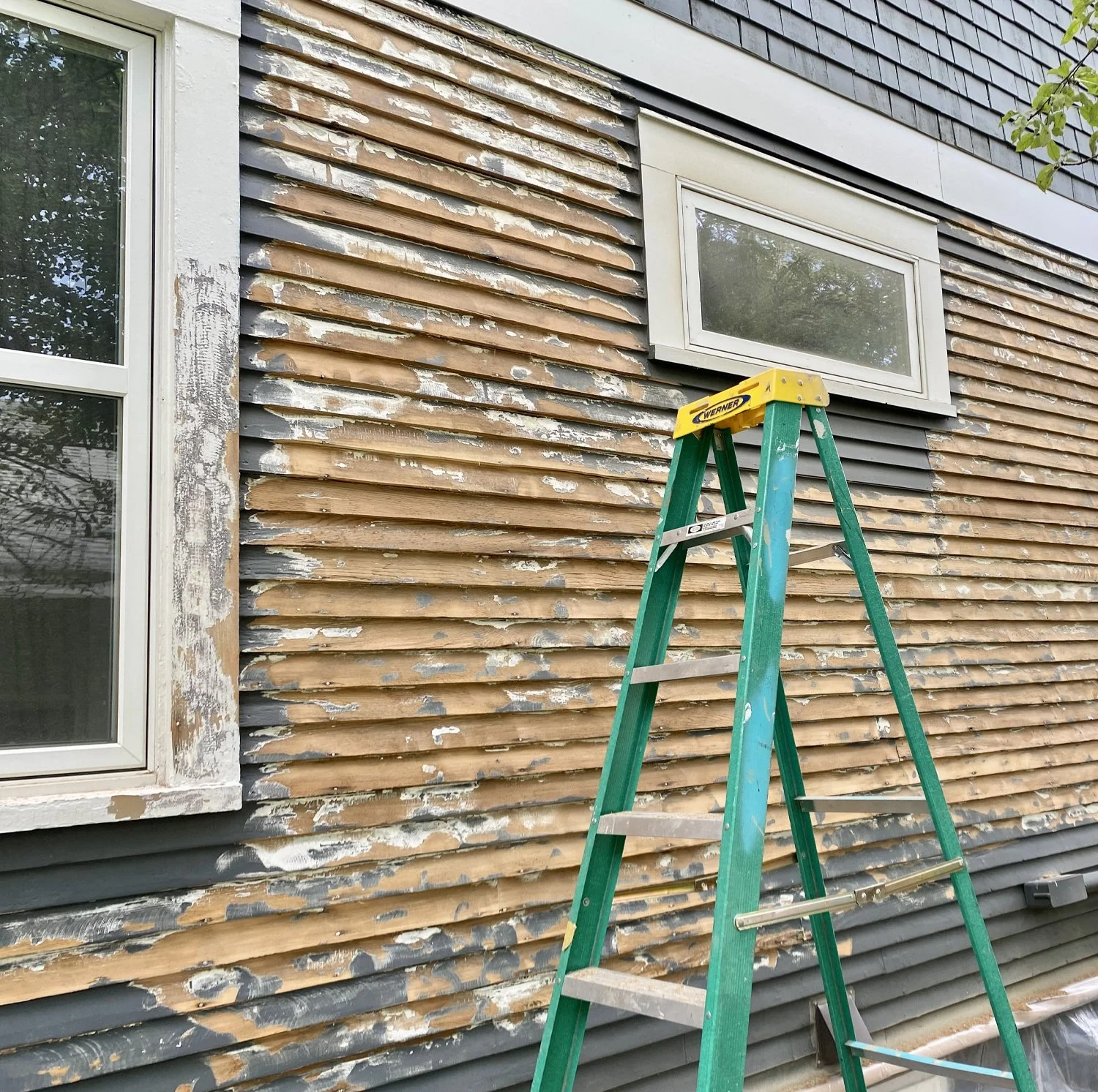 The exterior wall of a house with weathered horizontal wooden siding is being stripped for renovation. There are two white-framed windows visible, one larger with a transparent pane and a smaller one above. A green step ladder is positioned in front of the wall.