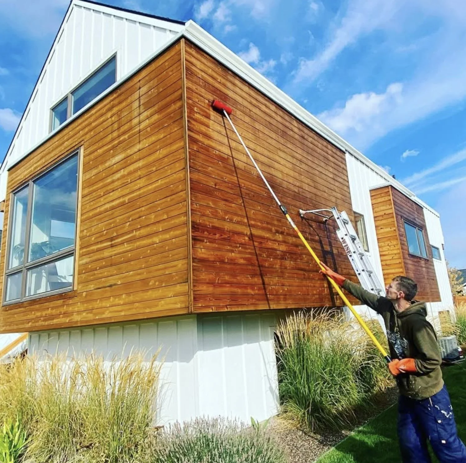 A person cleaning the exterior wooden siding of a modern house with a long pole and a power washer. The house has large windows and is elevated on a white foundation. The sky is partly cloudy with blue skies.