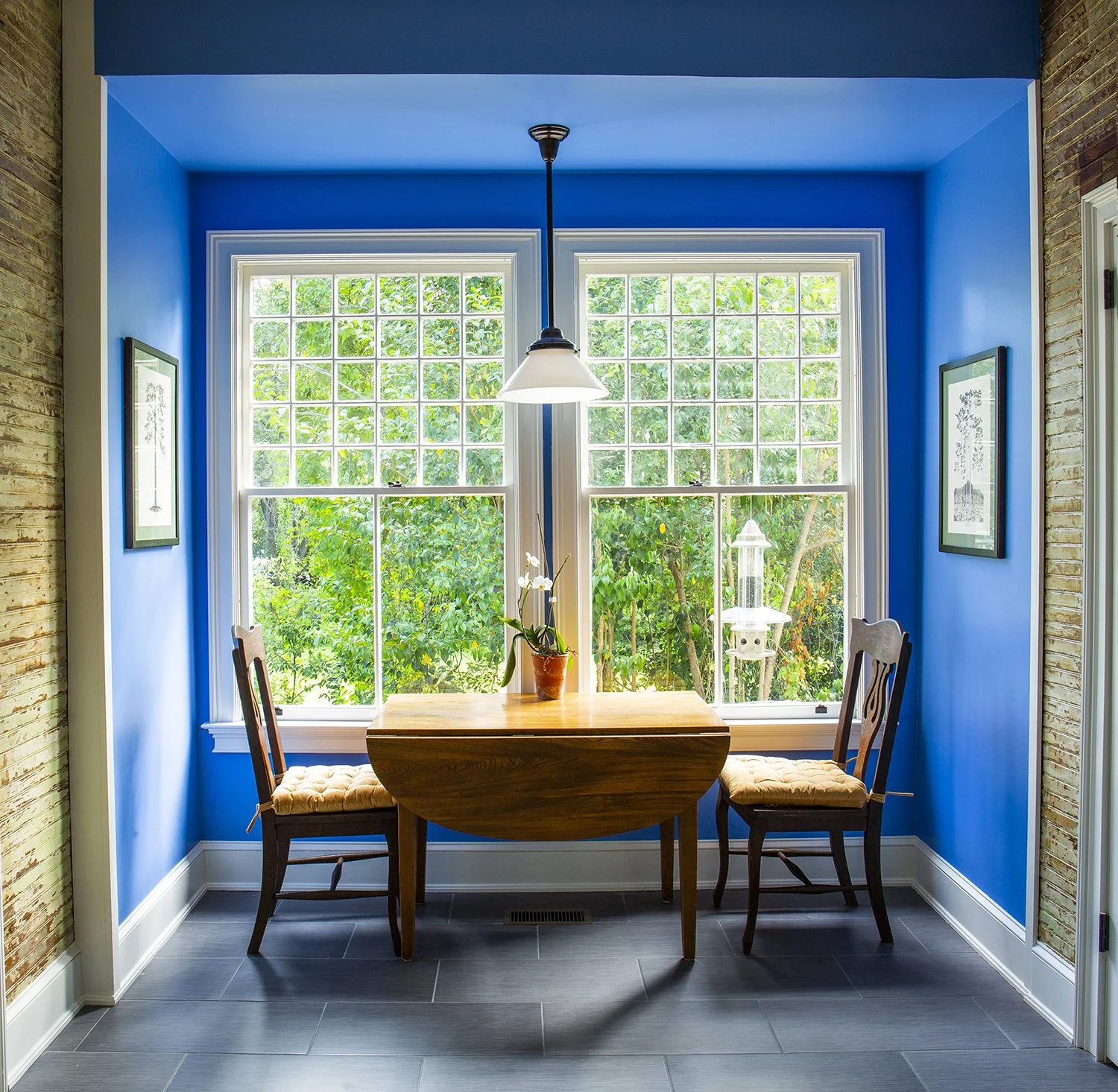 Dining nook with a round wooden table, two chairs with padded seats, a potted orchid, and a large window with a garden view, framed by bright blue walls and a hanging light fixture.