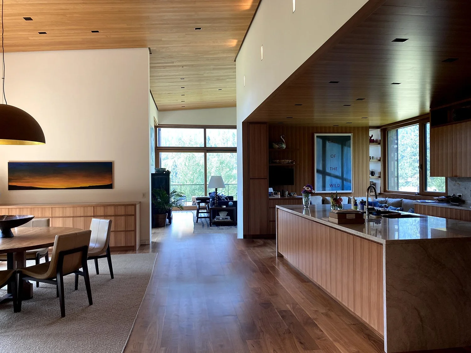 Open-concept living space with wooden ceiling and flooring, dining area with a round table and chairs, and a kitchen with a large island, windows with views of trees, and living area with furniture and a large window.