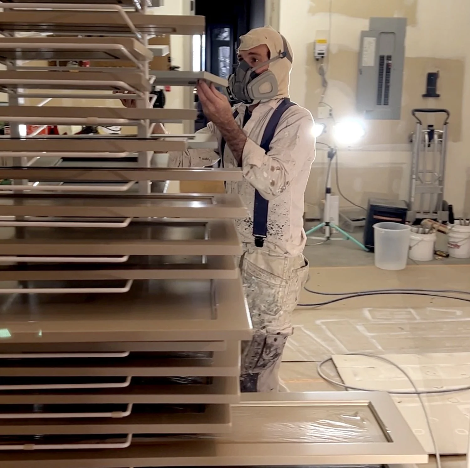 A person wearing a respirator mask, protective clothing, and suspenders working on wooden furniture or cabinetry in a workshop or construction site.