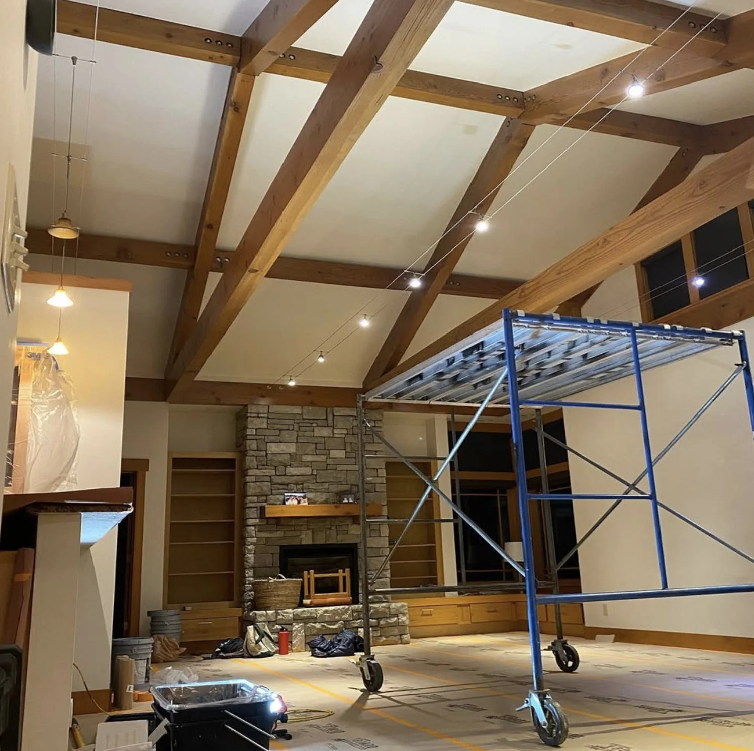 Interior of a room under construction with a high wooden ceiling, a stone fireplace, and a blue scaffold in the center. Construction tools and materials are scattered on the floor.