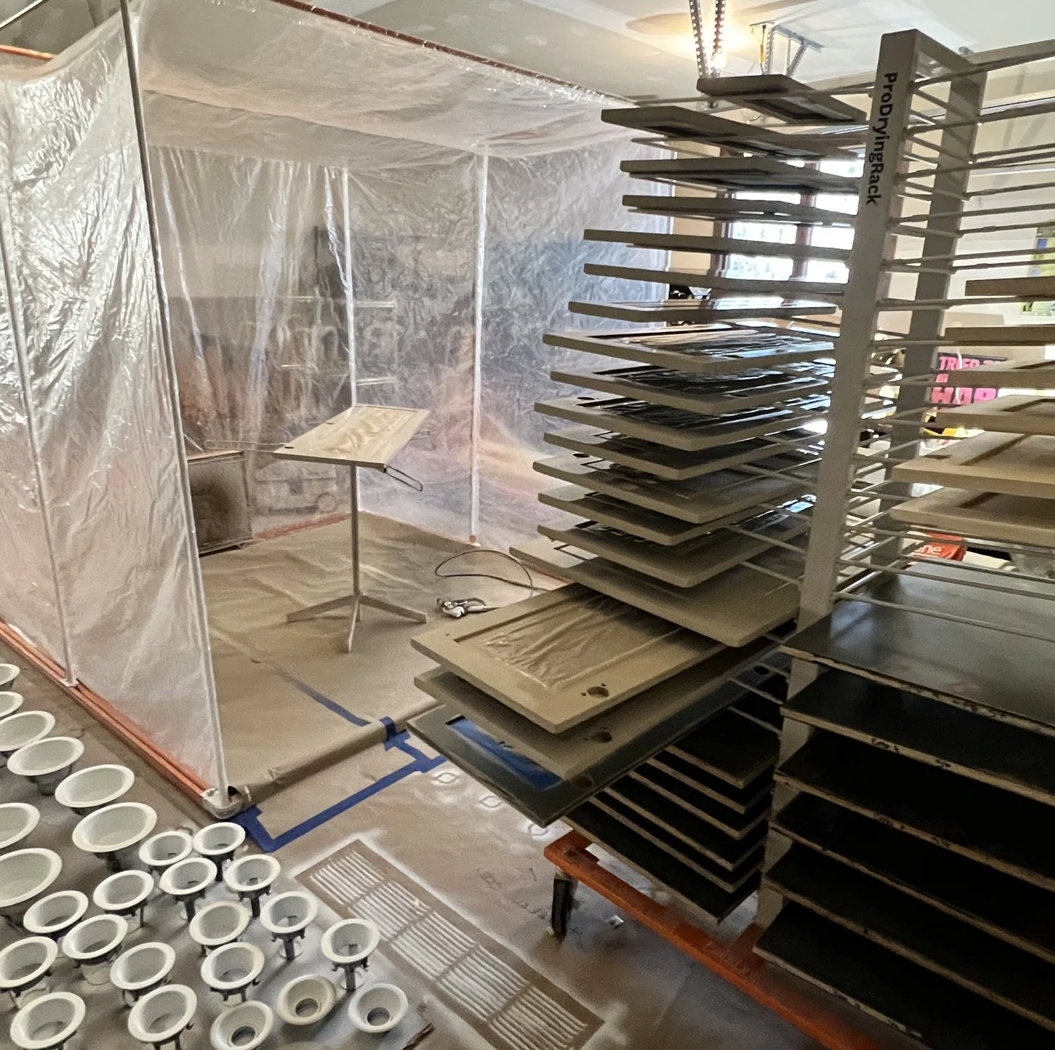 A painting or renovation workspace with plastic-covered walls and floor, a small stand with a black cord, and multiple shelves or trays stacked with panels or canvases, some wrapped in plastic.