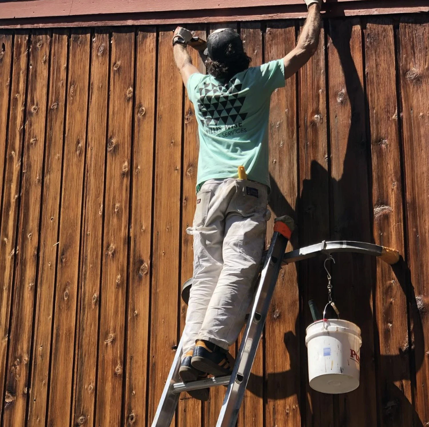 A worker standing on a ladder, painting or staining a wooden fence on a sunny day.
