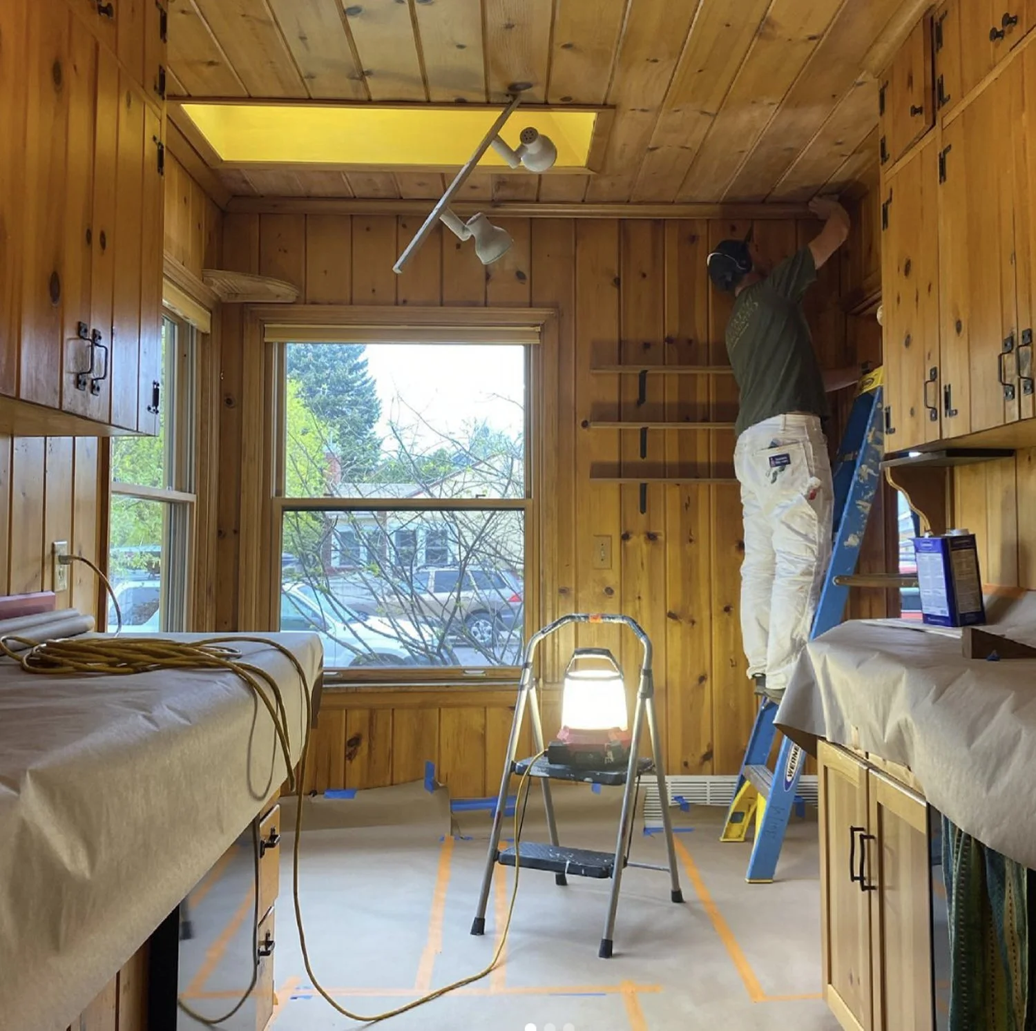A person standing on a ladder working on a wooden wall inside a room with wood paneling, a window, and a ceiling skylight, with tools and equipment around.