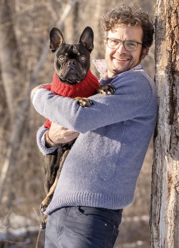 A man with curly hair and glasses smiling while leaning against a tree, holding a black French Bulldog wearing a red sweater.