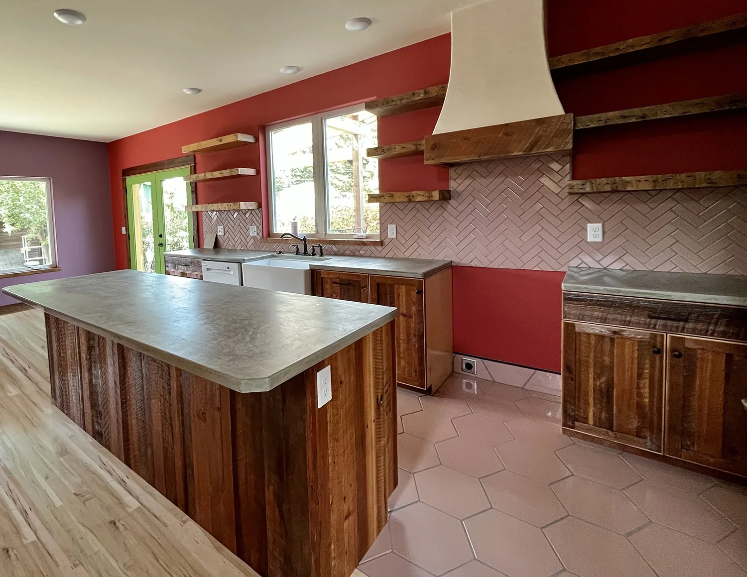 Kitchen with red and purple walls, wooden cabinets, a large central island with a stone countertop, a farmhouse sink, open wooden shelves, and a white range hood above a brick backsplash.