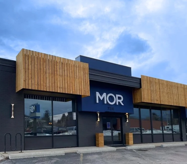 Exterior view of a modern commercial building with black walls, wooden accents, large glass windows, and a blue sign that reads 'MOR'.