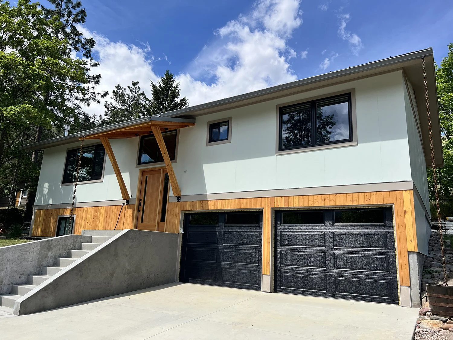 Modern two-story house with a concrete staircase and black garage doors, wooden accents, and large windows, surrounded by trees under a partly cloudy sky.
