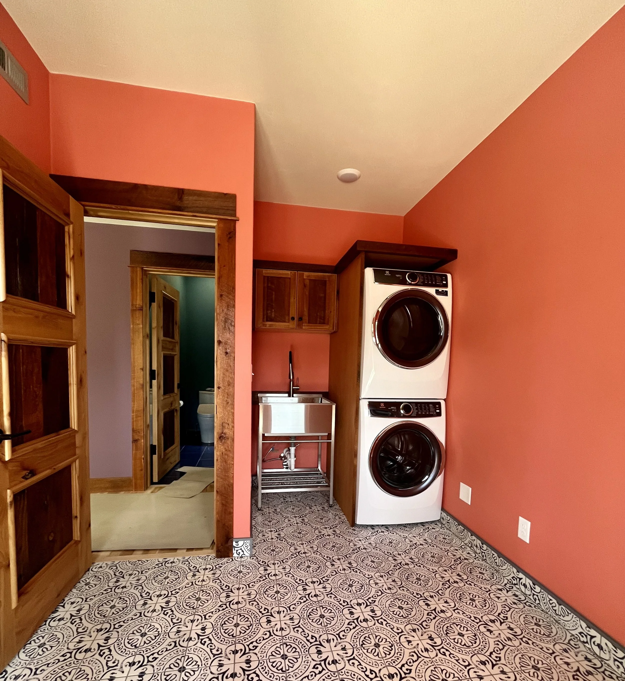 A laundry room with red walls, patterned black and white tile flooring, heated by a stacked washer and dryer, a utility sink, wooden cabinets, an open door leading to a bathroom with a toilet, and wooden door frames.